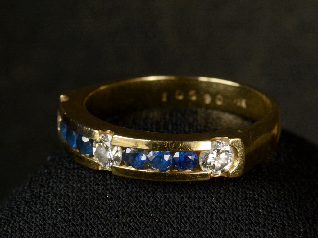 Elevated angled view of a vintage yellow gold ring with three diamonds interspersed with blue sapphires. Shown on white background. Faint markings inside the ring read "10590 18K"