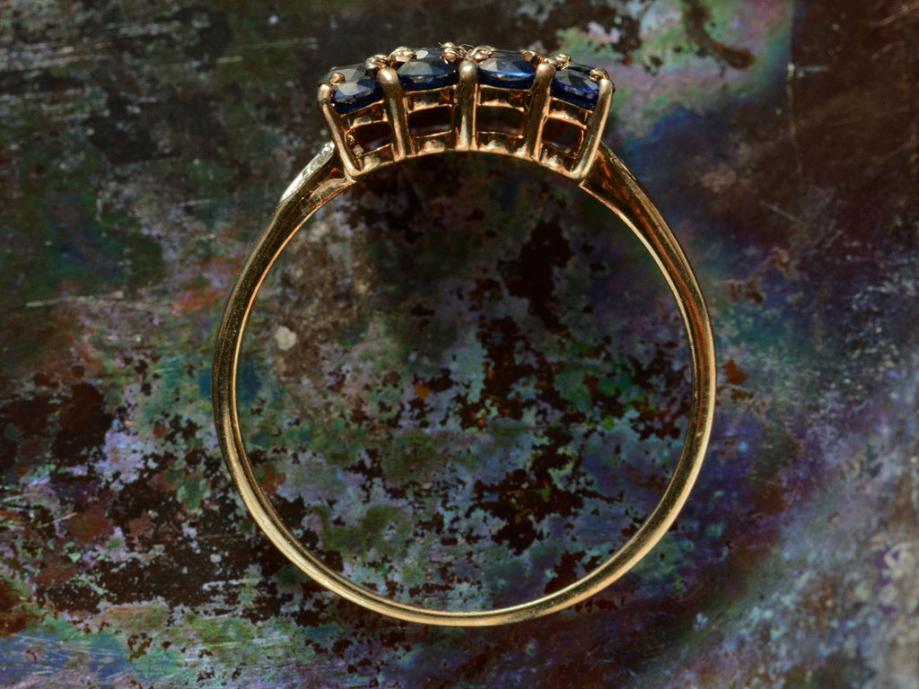 Side profile of a vintage yellow gold ring with a rectangular cluster of blue sapphires, shown on dark textured background.