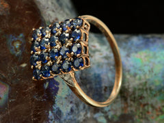 thumbnail of Angled view of a vintage yellow gold ring with a rectangular cluster of blue sapphires, shown on dark textured background.
