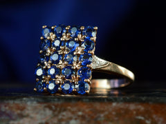 thumbnail of Angled front view of a vintage yellow gold ring with a rectangular cluster of blue sapphires, shown on dark textured background.
