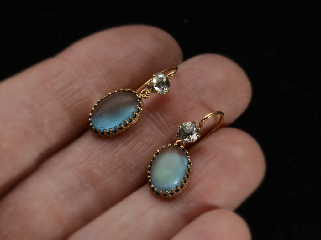 Pair of earrings with white crystal tops and oval pendants of pale blue glass with a reddish glow. Shown on  hand.