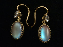 thumbnail of Angled view of earrings with white crystal tops and oval pendants of pale blue glass with a reddish glow. Shown on black background.
