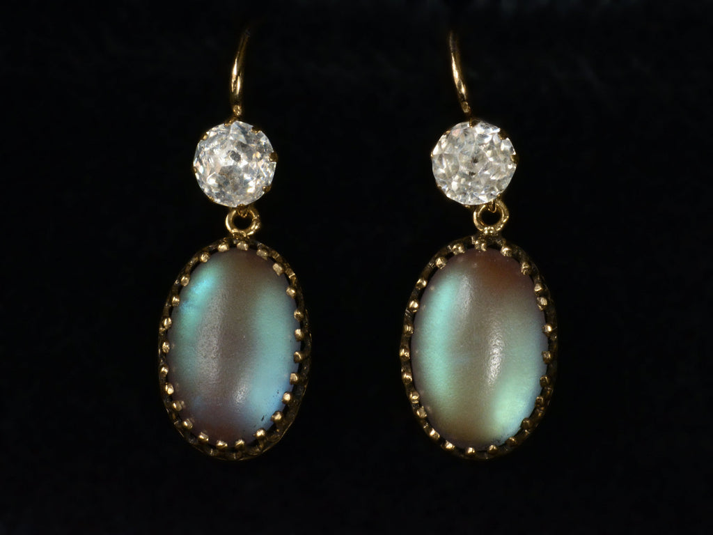 Pair of earrings with white crystal tops and oval pendants of pale blue glass with a reddish glow. Shown on black background.