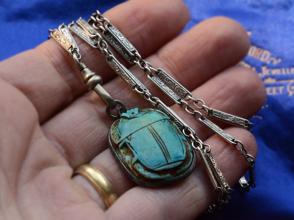 Hand holding turquoise-colored scarab pendant and Deco detailed silver chain.