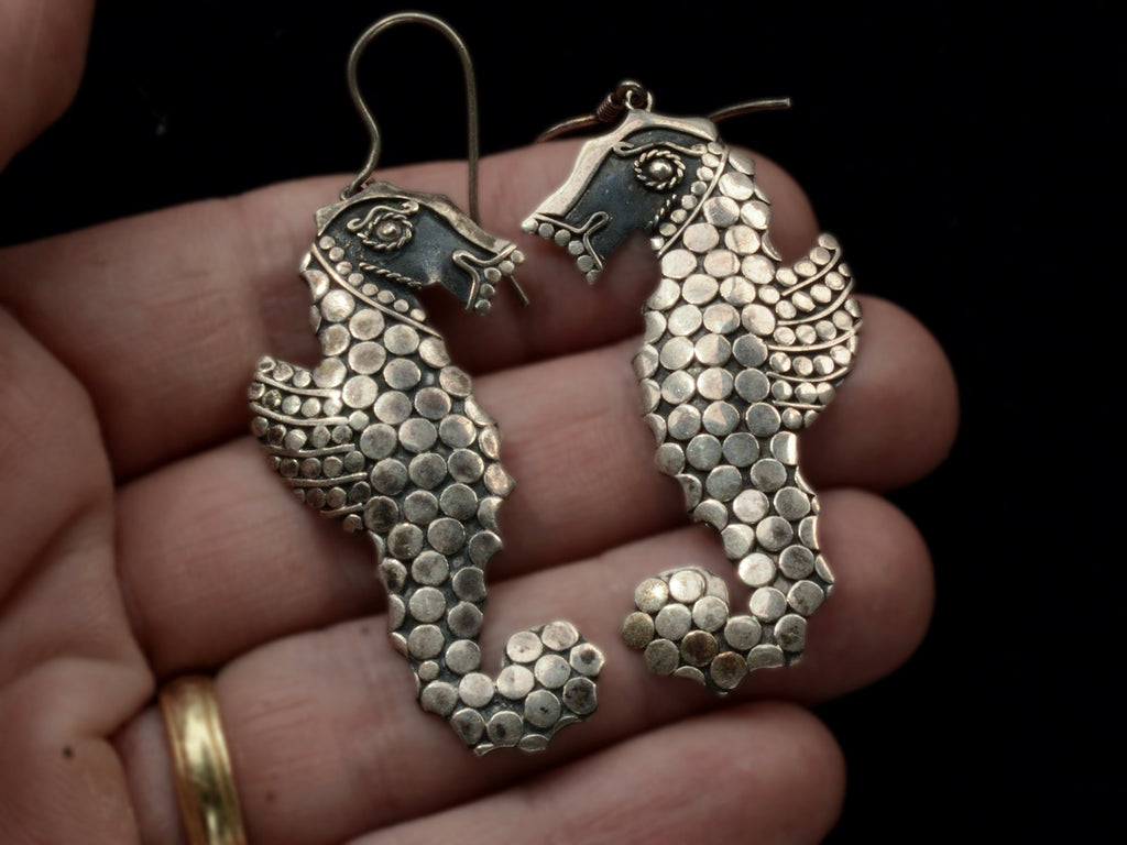 A pair of silver seahorse drop earrings textured with disks, shown held in hand.