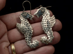 thumbnail of A pair of silver seahorse drop earrings textured with disks, shown held in hand.