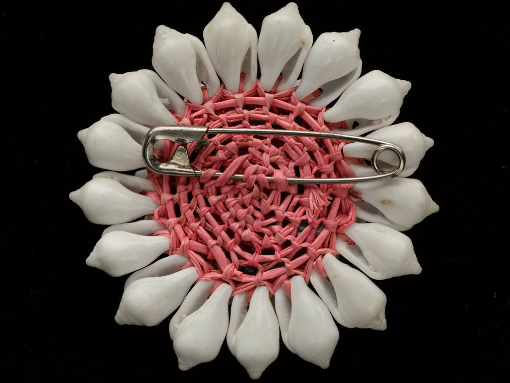 Reverse side of a vintage brooch featuring white shell petals arranged in a sunburst pattern around a central peach and brown shell cabochon, set in a pinkish-beige textured border, on black background. Shows safety pin fitting.