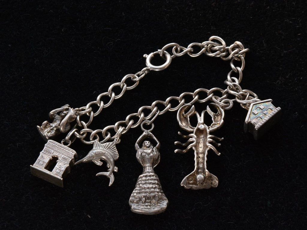 Reverse side detail of Vintage Silver Charm Bracelet showing a house, a lobster, a dancer, a fish, the Arc de Triomphe and a monkey. On a black background.