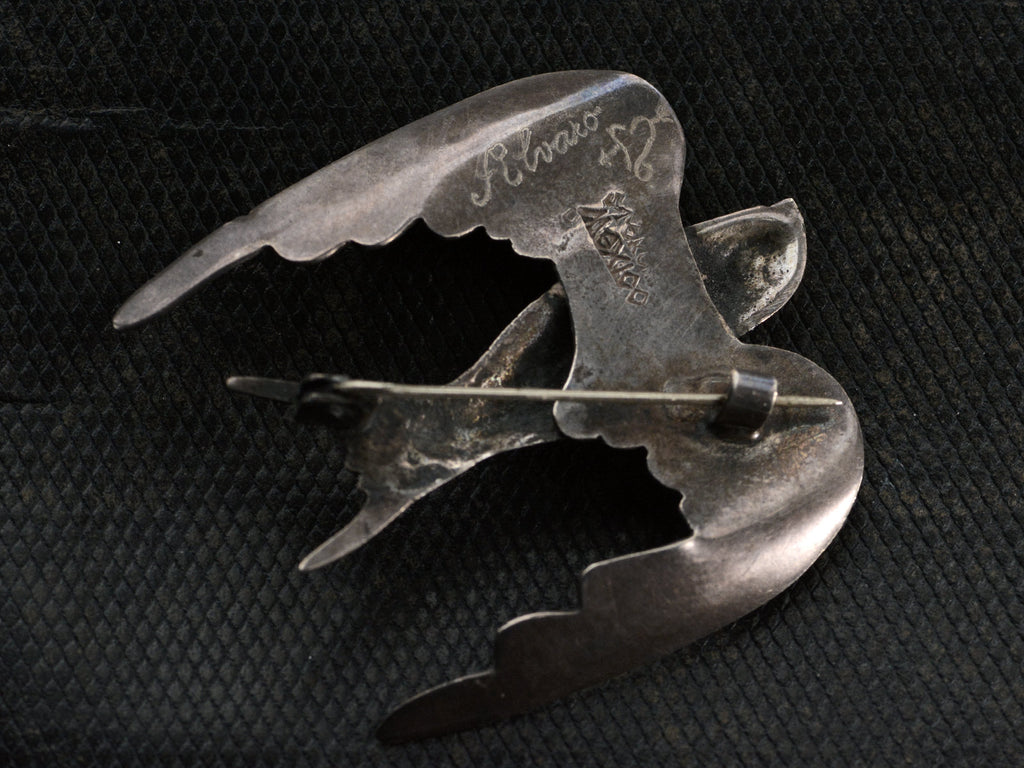 Reverse side of sterling silver dove brooch on dark textured background. Stamped "MEXICO" and signed "Alvaro" with a heart and arrow symbol.