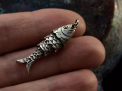 thumbnail of A small silver fish-shaped pendant with detailed scale engravings, held between a person's fingers for scale against a dark textured background.