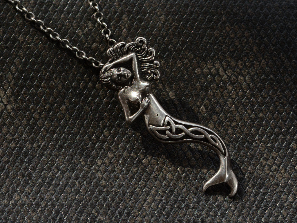 Detail of vintage silver mermaid pendant with Celtic design on tail, shown hanging from cable chain on dark textured background