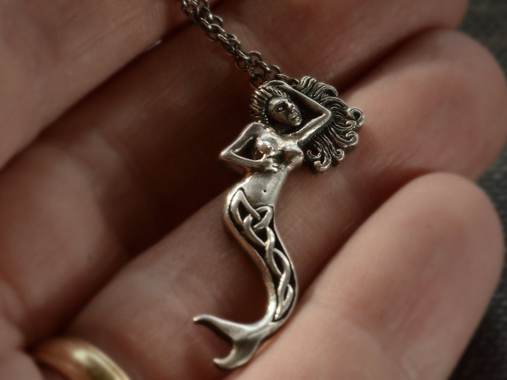 Detail of vintage silver mermaid pendant with Celtic design on tail, shown hanging from cable chain on hand