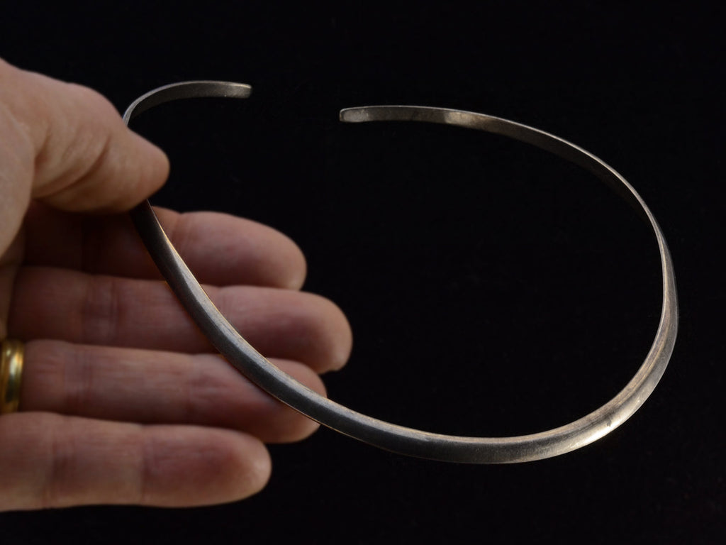 A hand holding a simple silver neck cuff against dark background.