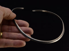thumbnail of A hand holding a simple silver neck cuff against dark background.
