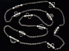 thumbnail of A c1990 long silver chain with interspersed tubular links, each decorated with pearls and a different geometric design or squiggle. Shown on black background.