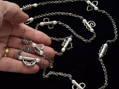 thumbnail of A c1990 long silver chain with interspersed tubular links, each decorated with pearls and a different geometric design or squiggle. Shown on held in hand against black background.