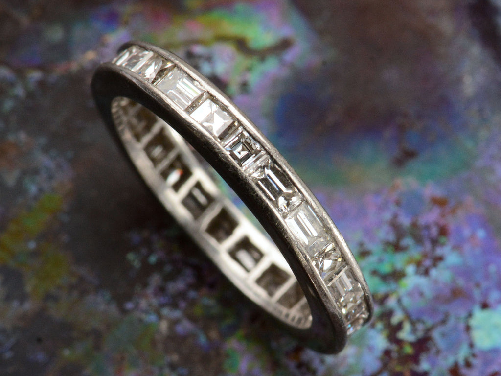 Detail view of a platinum eternity band set with square and rectangular diamonds, shown on dark colorful background.