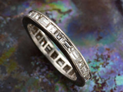 thumbnail of Detail view of a platinum eternity band set with square and rectangular diamonds, shown on dark colorful background.