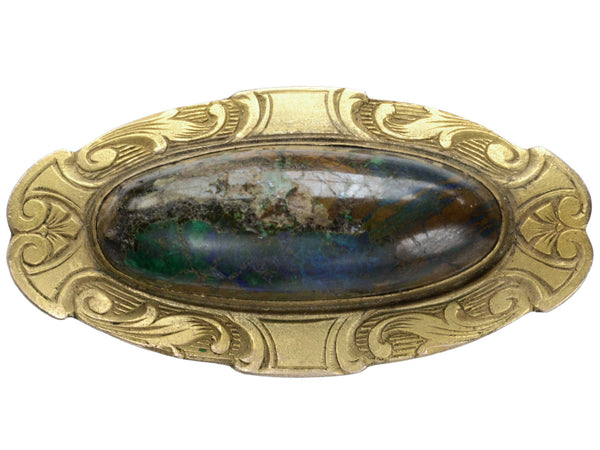 Oval brooch with a dark blue, green and rust colored cabochon set in a surround of decoratively carved gold plated brass, shown on white background.