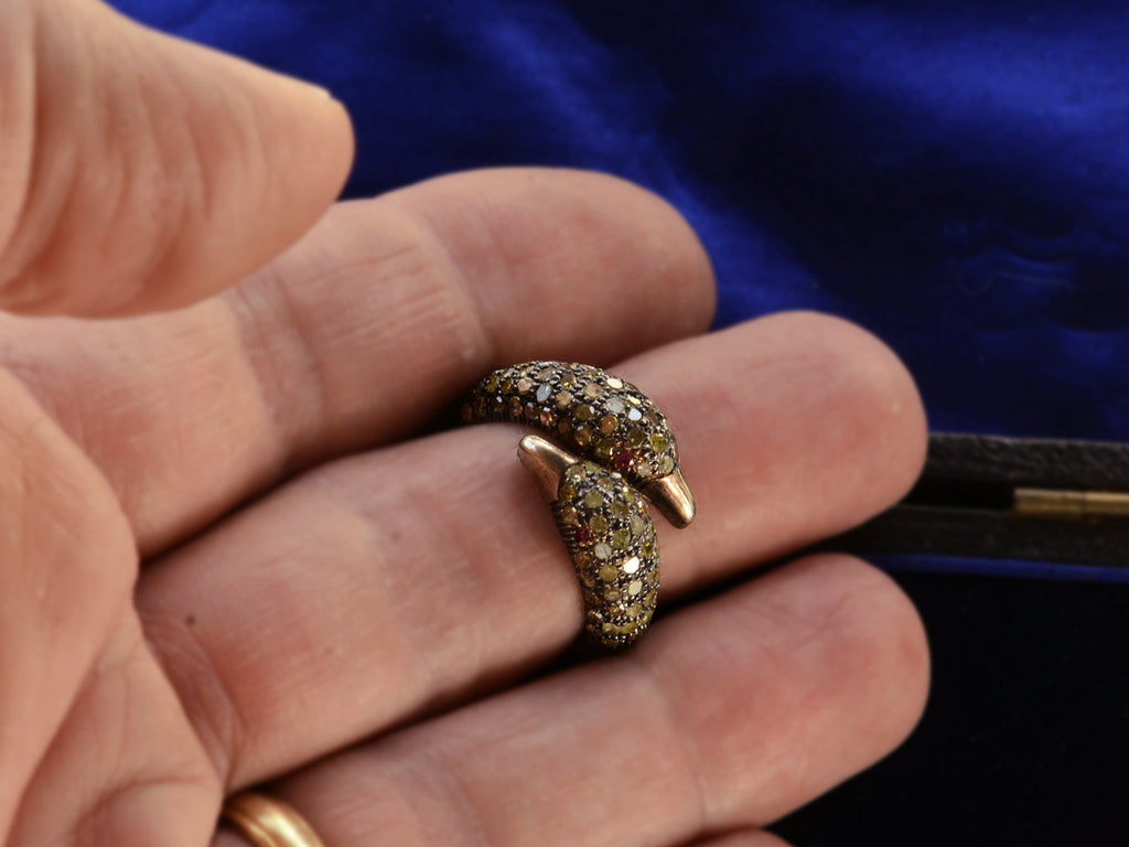 Figural ring with two swan heads, set with light yellow diamonds and ruby eyes in silver, shown on finger.