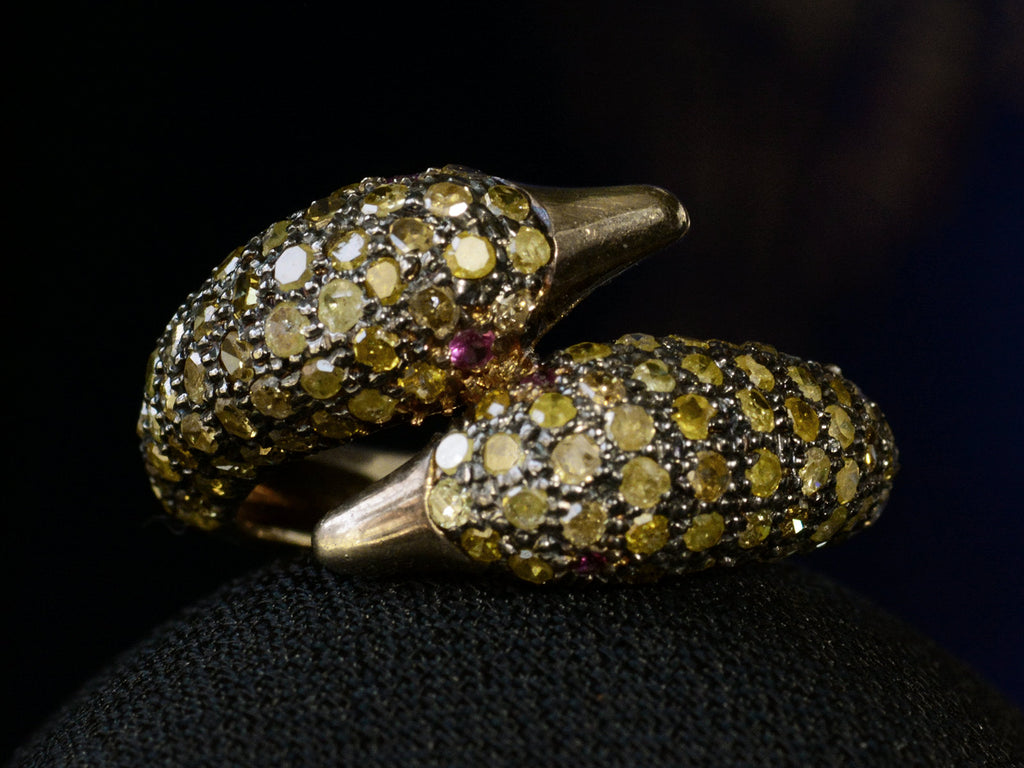 Front view of a figural ring with two swan heads, set with light yellow diamonds and ruby eyes in silver, shown on dark background.
