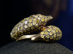 thumbnail of Angled view of a figural ring with two swan heads, set with light yellow diamonds and ruby eyes in silver, shown on dark background.
