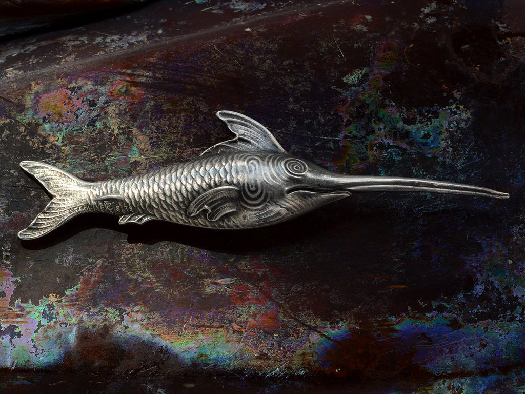 Backside of antique silver sewing stiletto in the form of a swordfish, shown on dark iridescent background. Stamps visible reading "STERLING" and "2179"
