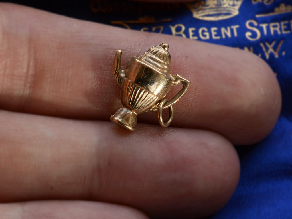 A gold 1964 Gold Teapot Charm shown on hand for scale.