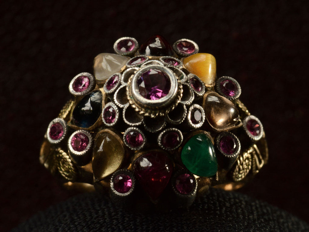Front view of tall tiered vintage ring set with rubies, and colorful gemstones in silver and gold. On dark background.