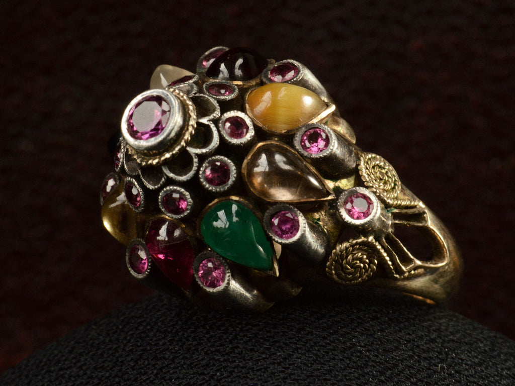Angled view of tall tiered vintage ring set with rubies, and colorful gemstones in silver and gold. On dark background.