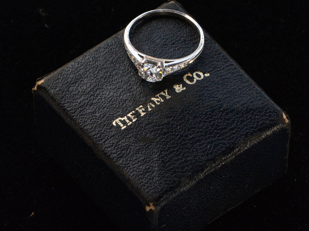 Elevated view of a c1930 Art Deco Tiffany & Co platinum engagement ring with a round old cut diamond and three smaller diamonds on each shoulder, shown sitting on top of its original "TIFFANY & CO" stamped ring box