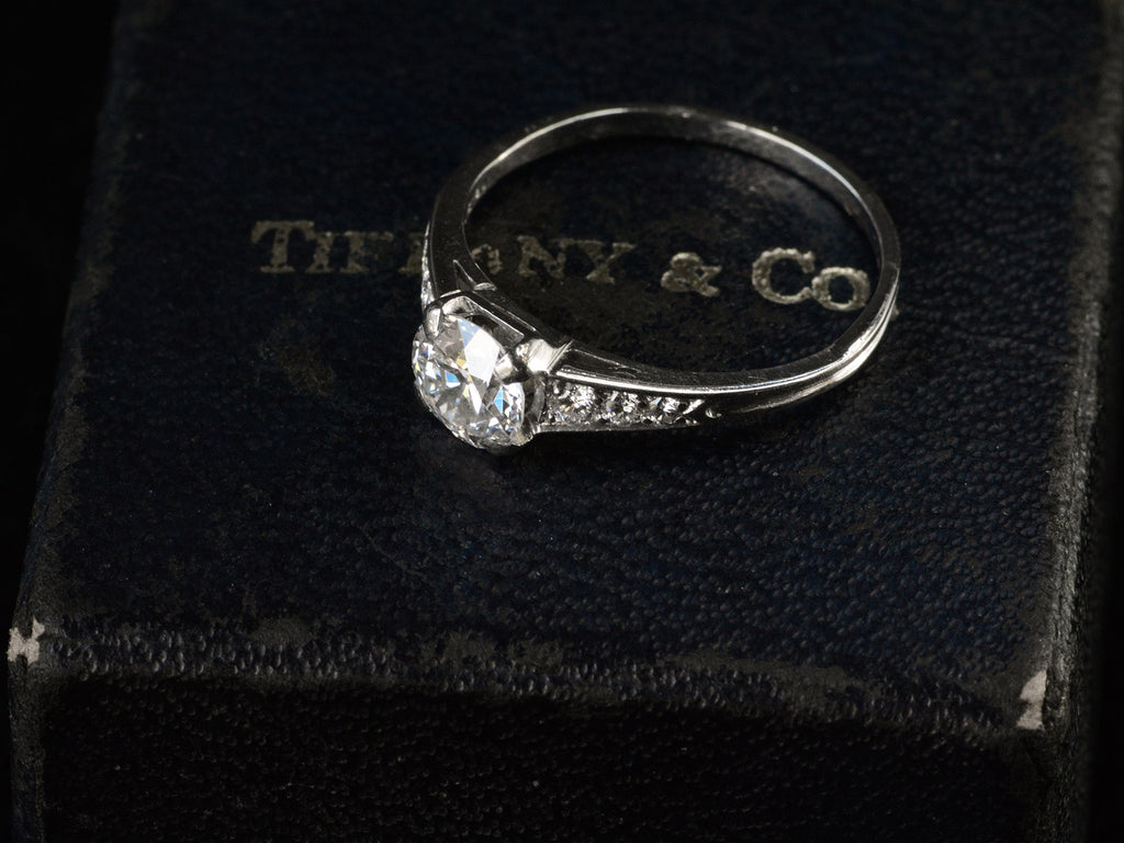 Angled view of a c1930 Art Deco Tiffany & Co platinum engagement ring with a round old cut diamond and three smaller diamonds on each shoulder, shown sitting on top of its original "TIFFANY & CO" stamped ring box
