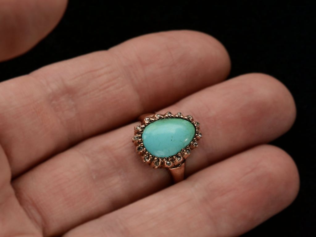 Antique rose gold ring with egg shaped turquoise surrounded by small rose cut diamonds, shown on hand.
