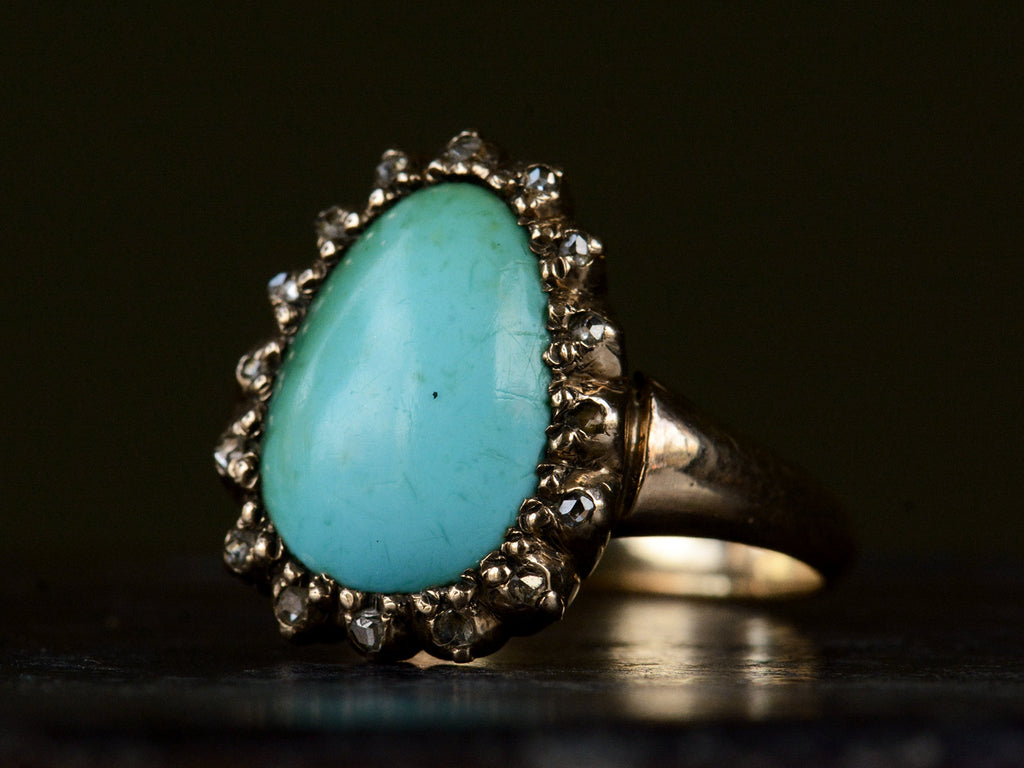 Angled view of antique rose gold ring with egg shaped turquoise surrounded by small rose cut diamonds, shown on dark background.