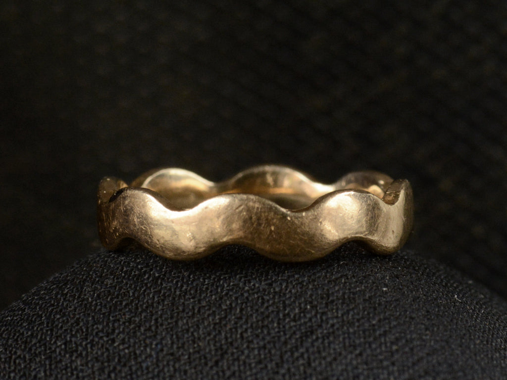Front view of a gold ring with a wave-form design, on dark background.