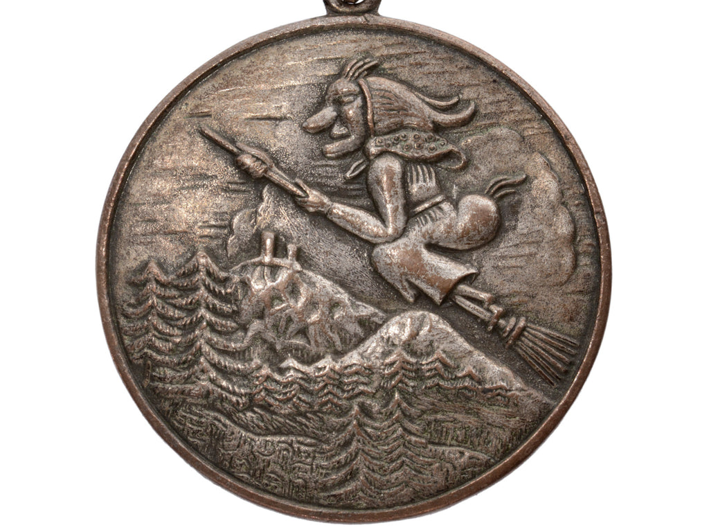 A silvered brass 1970s pendant with a witch flying on broomstick over mountains (on white background)