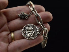 thumbnail of Hand holding a vintage sterling silver charm bracelet with charms depicting mating frogs and a pair of hares perched beneath a tree and a pair of crescent moons. Against black background.