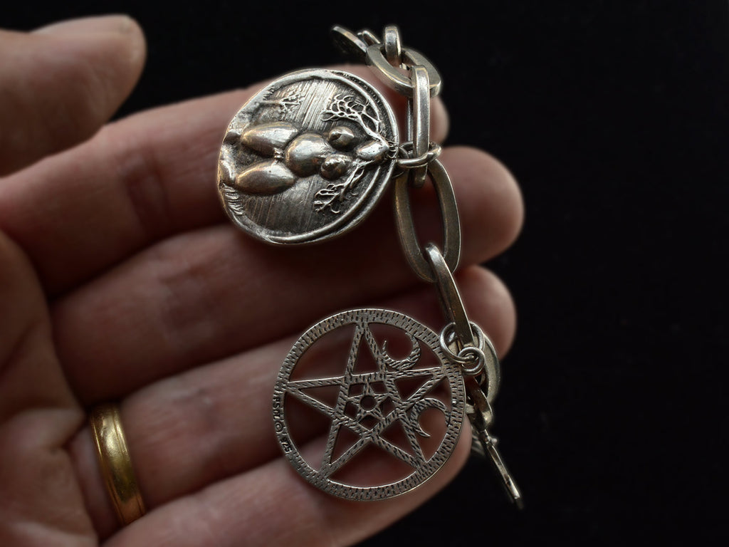 Hand holding a vintage sterling silver charm bracelet with charms depicting a nude woman and a pentagram. Against black background.