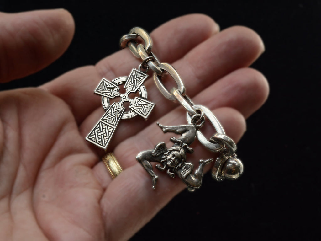 Hand holding a vintage sterling silver charm bracelet with charms depicting a Celtic cross and the Sicilian Trinacria. Against black background.